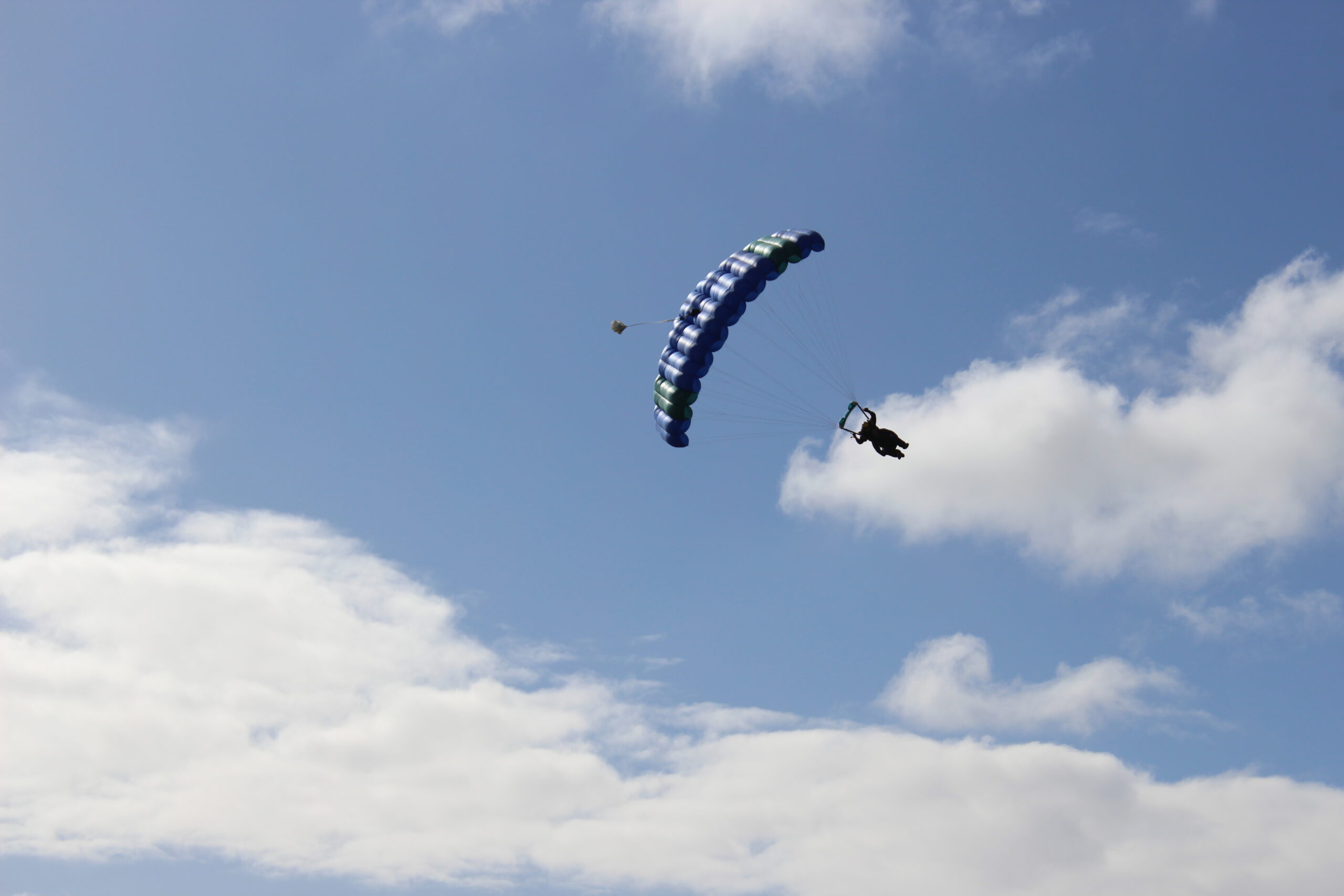 Two tandem skydivers descend under a blue and white canopy against a clear sky with scattered clouds.