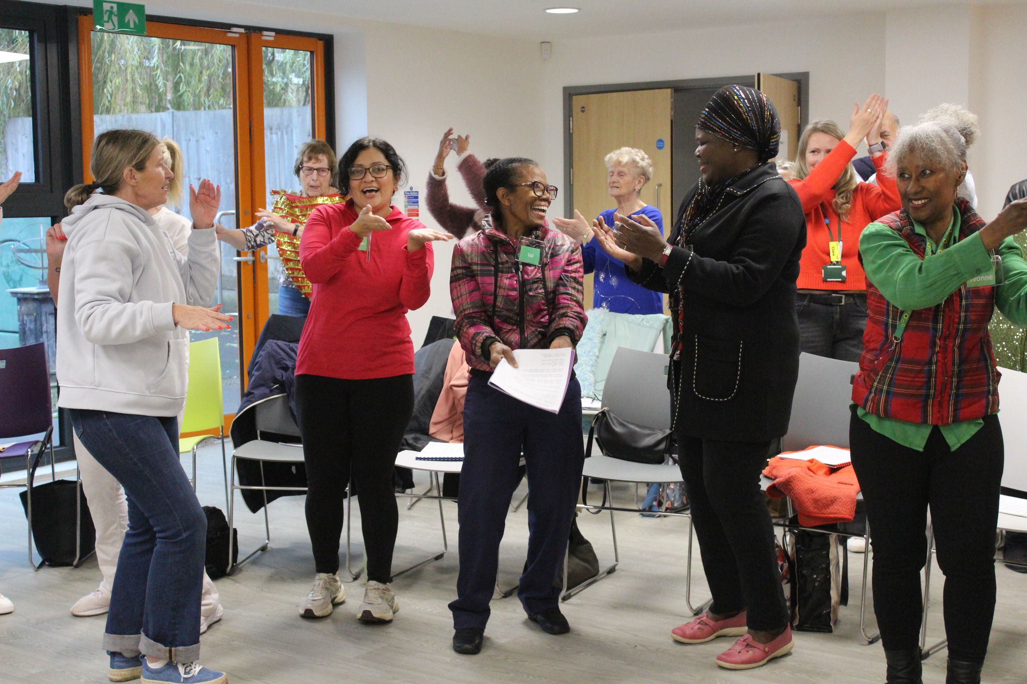 Diverse group of adults standing in a circle, clapping and smiling in a bright room.