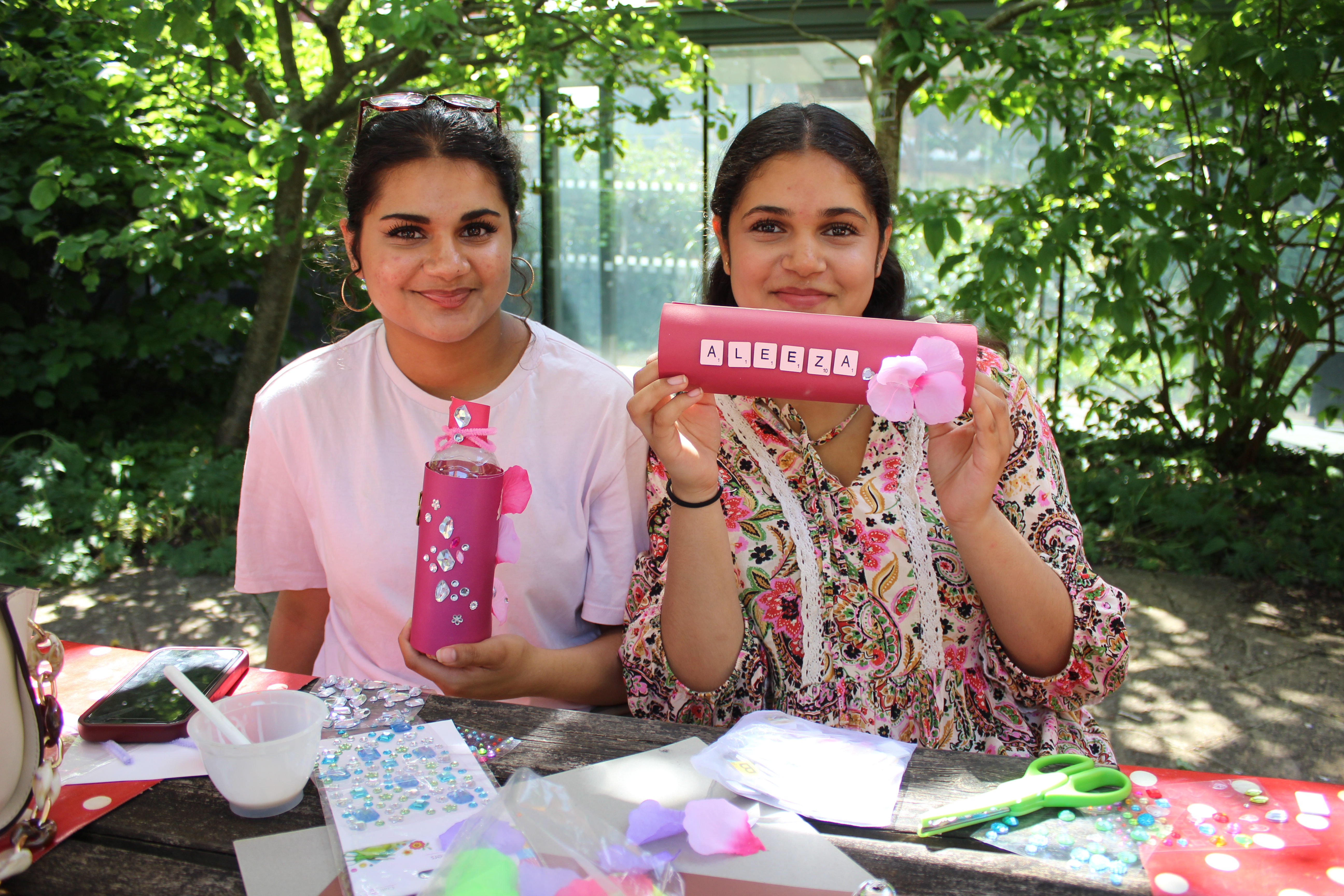 Two smiling young women craft outdoors at a table with pink decor; a pink case with letter tiles, gems and scissors, leafy backdrop.