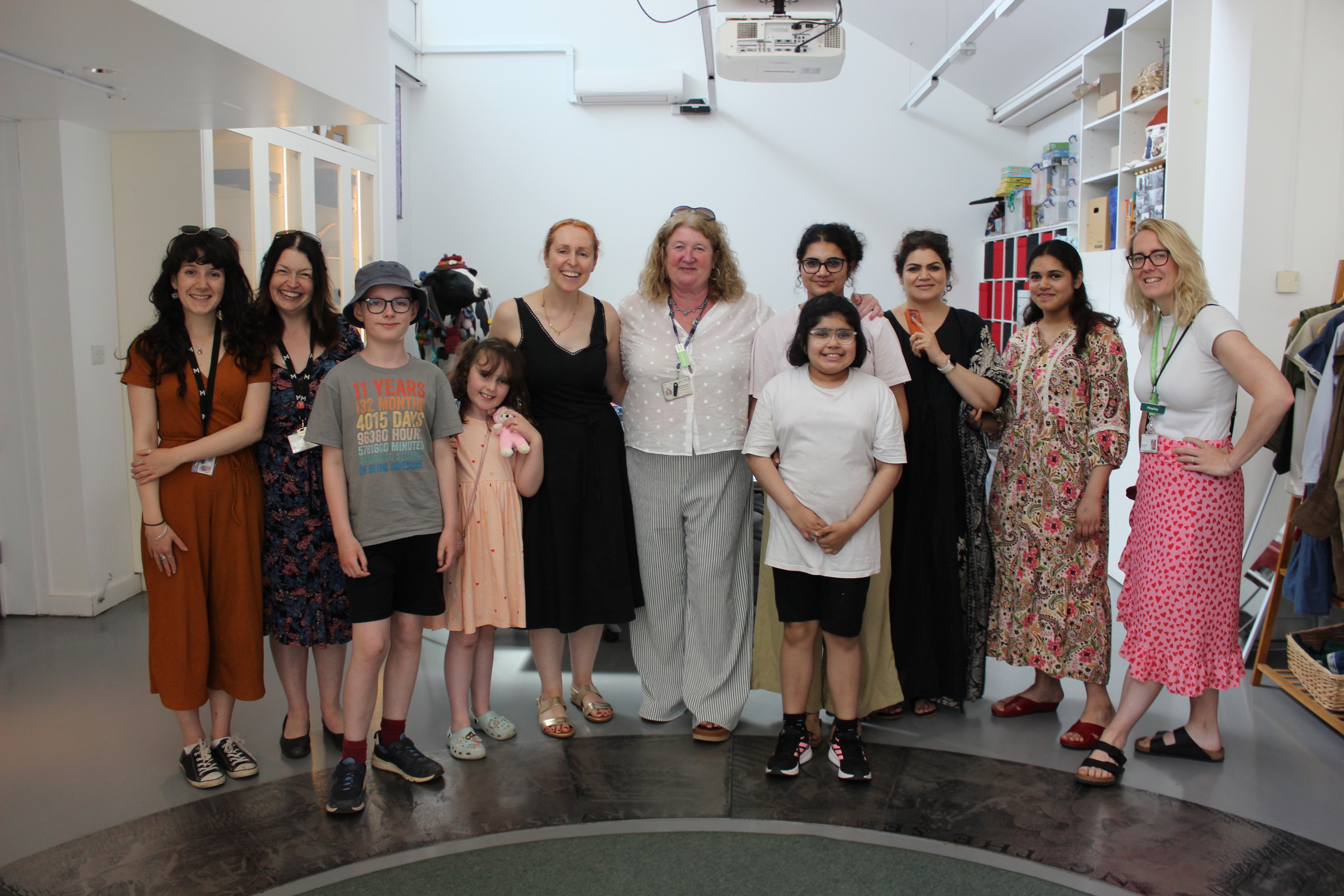 Smiling diverse group of adults and children stand in a bright room, row across the centre; white walls, shelving to the right, ceiling projector overhead.