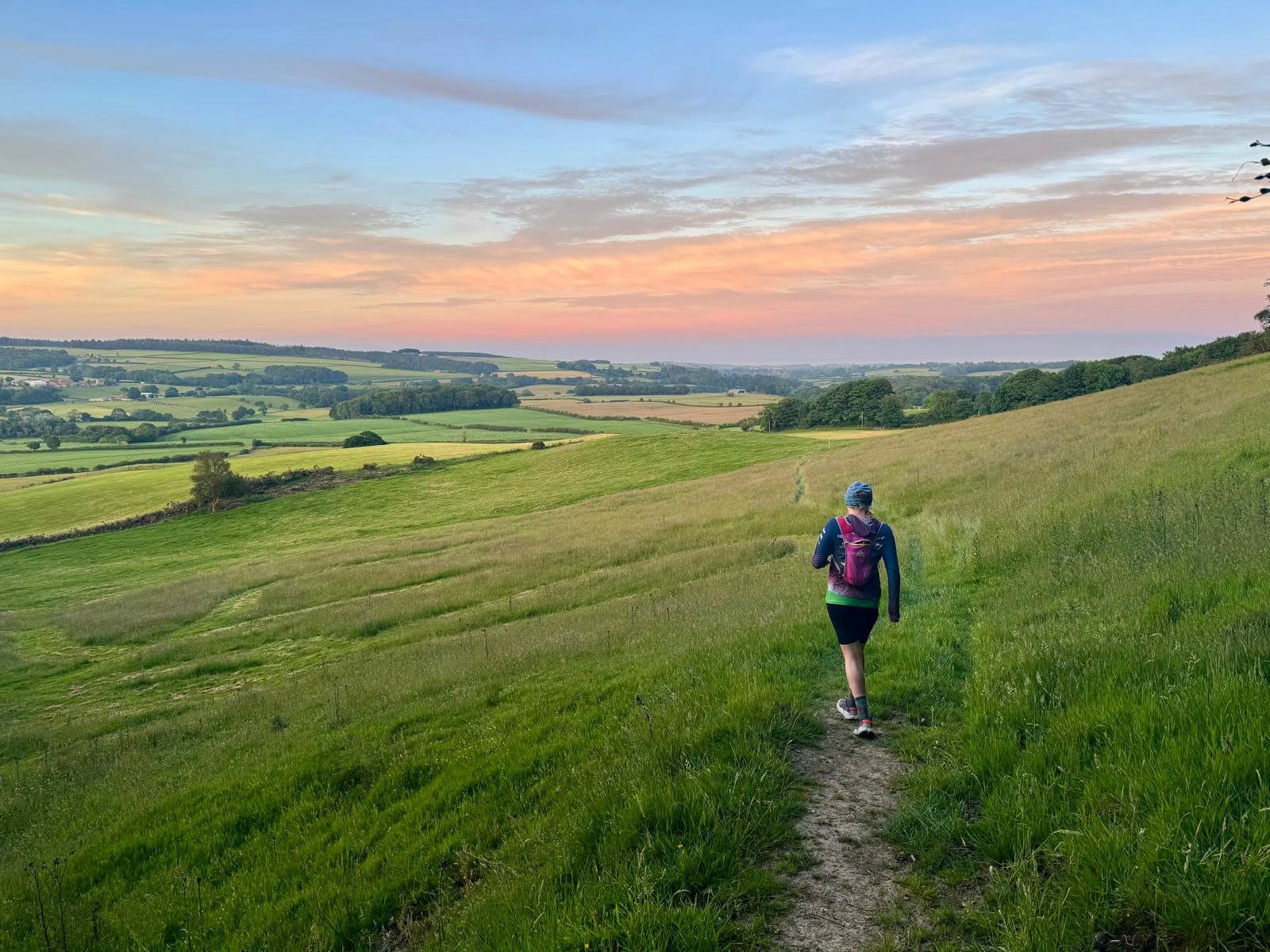 Hiker with purple backpack walking along a grassy hillside trail at sunset over rolling countryside.
