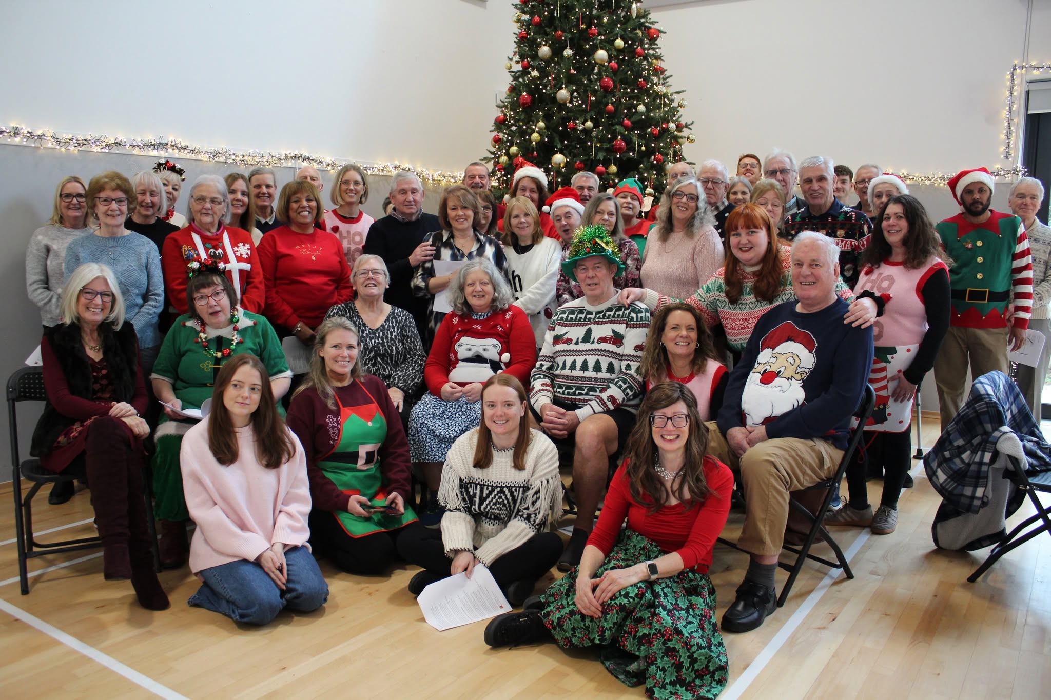 Group of people in Christmas jumpers and hats posing for a festive photo in a hall with a decorated Christmas tree.